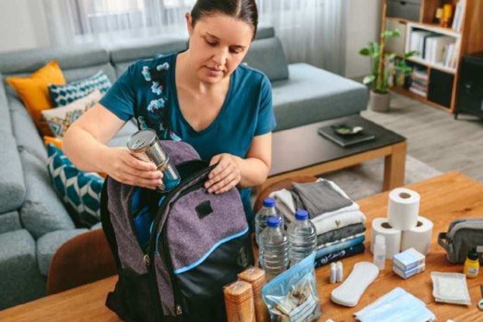 Woman putting cans of food to prepare emergency backpack in living room