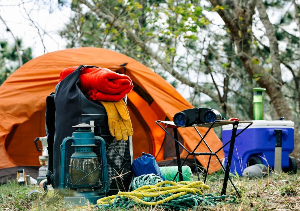 A tent surrounded by essential survival gear, including blankets, rope, and a cooler box, set up in a forest clearing.