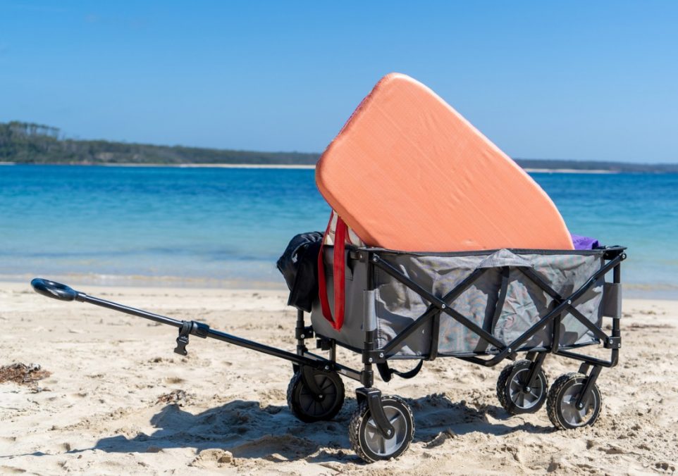 A gray beach wagon on the beach holds an orange boogie board, towels, and beach toys.