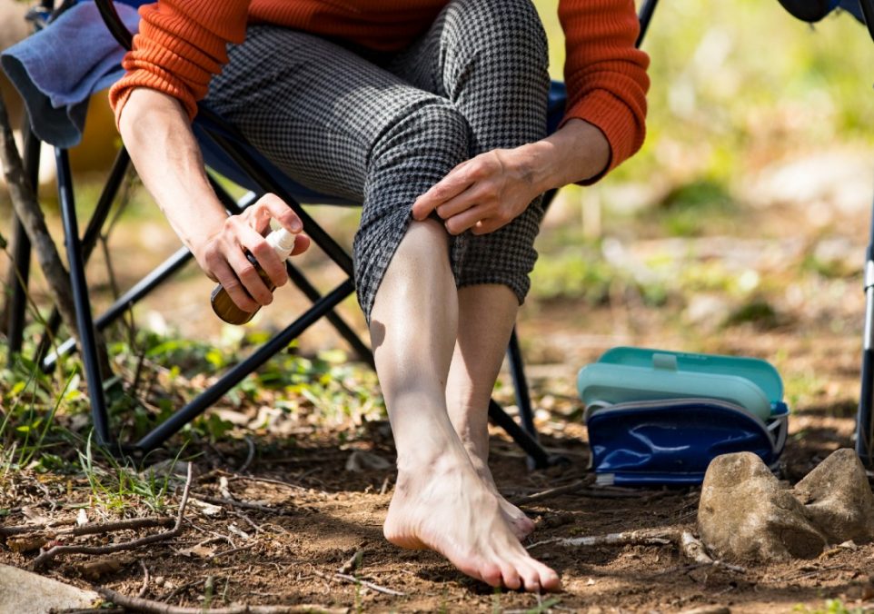 A woman spraying tick repellent on her leg as she sits on a camping chair.