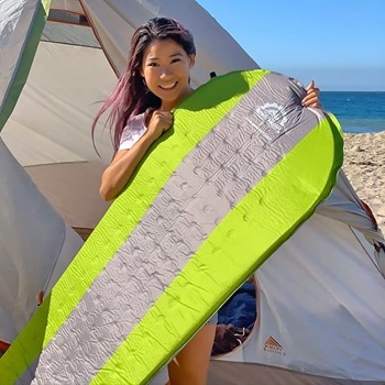 A pretty woman holding the Gear Doctors Camping Sleeping Pad on a beach with a tent behind her.