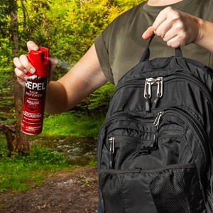 A man sprays a black backpack with REPEL Tick Repellent, ensuring protection against ticks while he's outdoors.